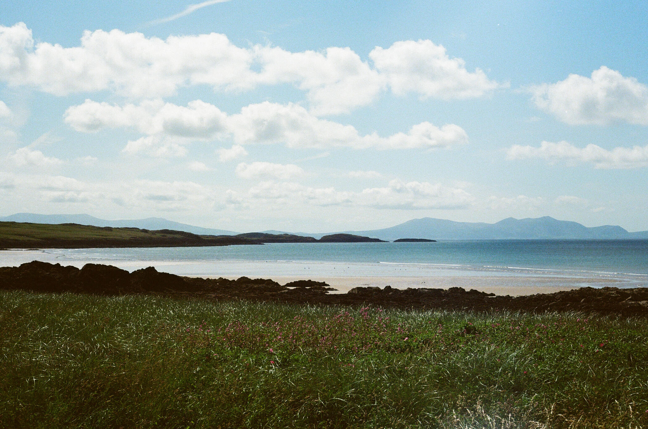 An image of a blue sky with white fluffy clouds, hills on the horizon, a blue sea, and grass in the foreground.